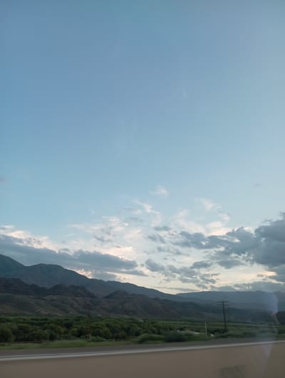 The image depicts a landscape scene in Kok-Moinok - 1, Kyrgyzstan, likely captured from a moving vehicle. The perspective is a wide shot with the camera oriented vertically. The left two-thirds of the image are dominated by a clear blue sky with scattered white and grey clouds, suggesting it is daytime, possibly late afternoon or early evening given the soft lighting on the clouds.
The right third of the image reveals a mountainous terrain. The mountains are layered and appear arid, with some vegetation visible in the lower valleys and on some slopes. The greenery suggests a fertile area within the mountainous landscape. The sky extends beyond the mountains, and the clouds are more prominent towards the right edge.
In the middle ground, a power line with two crossbars stands vertically against the backdrop of the mountains and sky. A second, shorter pole is visible to its right. These elements indicate some human infrastructure present in the otherwise natural setting. The overall mood is serene and expansive, capturing the vastness of the sky and the rugged beauty of the mountains. There is no visible text in the image.