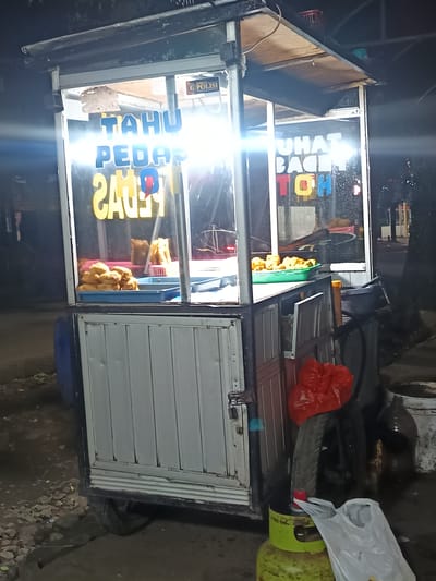 This media file depicts a street food stall at night, likely in Karangtengah, Indonesia. The stall is illuminated by bright lights, highlighting the food and signage. The primary subject is the food cart itself, which is filled with fried food items, possibly tofu or similar snacks, displayed in trays.
Visible text on the stall reads "TAHU PEDAS" which translates to "Spicy Tofu", suggesting the type of food sold. Another sign above the display reads "UHA BACOH TOH", which is less clear in its meaning without further context, but might be a brand name or a slogan. A small "POLISI" (Police) sticker is also visible on the upper part of the stall.
The scene is set outdoors, on a paved or dirt ground, with some debris and stones visible. In the background, trees and what appears to be a building or fence can be seen, along with some street lighting. To the side of the stall, there is a gas cylinder, a spare tire with a red plastic bag on it, and other miscellaneous items, indicating it is a functional street vending setup.
The time of day is clearly night, given the darkness of the surroundings and the artificial lighting of the stall. The weather appears to be calm, with no indications of rain or strong wind. No people are visible in the frame, so the activity depicted is the stationary operation of the food stall. The overall mood is one of a typical, bustling street food scene at night.