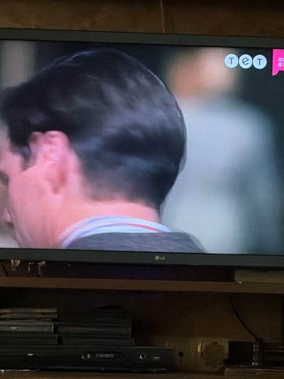 The image captures a close-up view of a television screen displaying a person's head and shoulder, with a visible LG logo at the bottom center of the screen. The background of the television shows a blurred, light-colored scene, possibly an interior setting, with three circular logos in the upper right corner reading "TET". Adjacent to these logos is a pink rectangular graphic. The television is placed on a wooden shelf, beneath which is a black DVD player and a stack of what appear to be CD or DVD cases. The scene is dimly lit, suggesting it is indoors. The location in the city of Osolynka, Ukraine, is not discernible from the image itself.