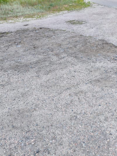 The image shows a close-up view of a gravel road with patches of grass growing on the sides. The road surface is uneven, with small stones and dirt visible. There are also some darker patches that appear to be damp or discolored. In the background, there is more grass and vegetation, suggesting a rural or undeveloped area. The lighting suggests it is daytime, and the weather appears to be clear. There is no visible text in the image. The scene is generic and does not depict any specific activity or event. The location is identified as Matiashivka, Ukraine, which could imply a rural setting within that region.