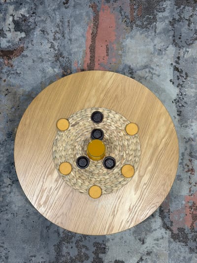 The image displays a close-up, overhead view of a round wooden table with a woven placemat in the center. The placemat holds several dark, circular game pieces and some small, round, yellow objects, possibly cookies or candies. A small glass filled with a yellow liquid, likely juice, sits on the placemat. The table is placed on a rug with an abstract, textured pattern in shades of gray, blue, and rust. The scene appears to be indoors, possibly in a home or cafe, and there are no people visible. The specific location of this scene within Sosnowiec, Poland, cannot be determined from the provided image.