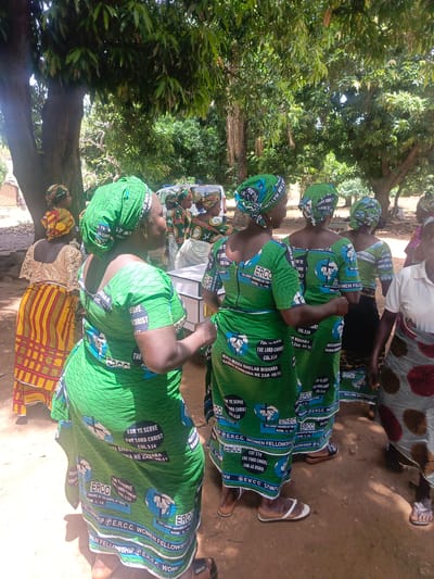 A group of women stand together in an outdoor area, likely in Ninzo South, Nigeria. The women, many dressed in matching green outfits, with the inscription "E.R.C.C. WOMEN FELLOWSHIP" and the phrase "FOR YE SERVE THE LORD CHRIST COL.3:24" printed on them. They are under the shade of trees. A few women are wearing different patterns. A white ambulance can be seen in the background. The ground appears to be dirt.