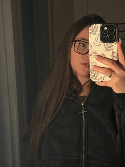 The scene depicts a mirror selfie of a female individual in an indoor setting within the city of Prikubanskiy, Russia. The subject, with long, dark brown hair, is wearing dark-rimmed rectangular glasses, a dark, zippered hoodie, and a metallic four-pointed star or cross pendant hanging from the zipper. A simple metallic band is visible on her left ring finger. She holds a smartphone, likely an iPhone with a multi-lens camera array, in her right hand, partially obscuring the lower half of her face. The phone is encased in a light-colored, patterned case featuring vertical stripes and subtle floral and butterfly designs in muted tones. Her fingernails are manicured with a light polish. The background, reflected in the mirror, is dimly lit and consists of indistinct vertical elements, possibly part of a doorframe or wall.