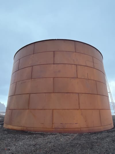 A large, cylindrical tank made of riveted metal plates dominates the frame. The tank's surface has a reddish-brown hue and shows signs of aging or oxidation. The surrounding environment includes a gray sky and a rough, rocky ground or embankment. A metallic structure is visible in the background.