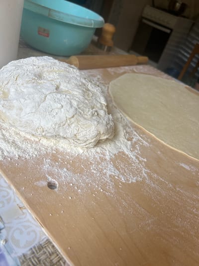 The image shows a close-up of dough and a flattened piece of dough on a wooden surface. A wooden rolling pin is visible in the background, along with a light blue bowl. The scene appears to be in a kitchen environment, with a stove and chair visible in the background. The lighting suggests an indoor setting. There are no people visible in the image. The location in the city of Staraya Sal'ya, Russia is not discernible from the image itself.