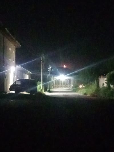 The image depicts a dark, outdoor scene, likely in the city of Shar-Bakr, Uzbekistan. A dark-colored van is parked on a paved surface next to a building on the left. The building has lit windows. Further down the paved area, a large gate or garage door is visible, illuminated by lights. Trees and vegetation are present on the right side of the image, and a faint crescent moon is visible in the dark sky. The overall atmosphere is dim and appears to be nighttime.