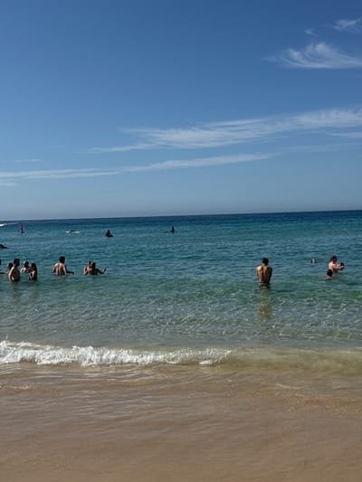 A beach scene features a clear blue sky with faint clouds. A line of people are swimming in the ocean near the shore. The water is clear and reflects the sky. A sandy beach extends to the right, marked by white waves rolling in. The scene is captured from a high angle, lending a broad perspective to the tranquil coastal setting.