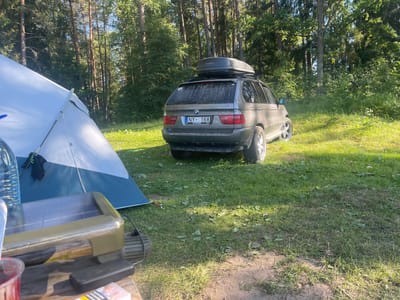 The image depicts a campsite in a forest clearing near Lielstraupe, Latvia. A grey BMW X5 SUV, equipped with a roof box, is parked on the grass. A partially erected blue and white tent is situated to the left of the vehicle. In the foreground, a wooden table holds a portable solar panel, a water bottle, and a pack of cigarettes. The scene suggests a camping trip or outdoor excursion. The sun is shining brightly, casting shadows on the grass, indicating it is daytime. The surrounding trees suggest a wooded or forested area. The license plate of the car reads "NY 168". A sticker on the rear window of the car says "UMBRELLA". The text "N-RODRIVE" is visible on the roof box. The overall mood is peaceful and serene, characteristic of a nature retreat.