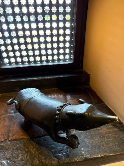 A high-angle shot shows a metal sculpture of an animal, possibly a dog or ox, mounted on a brick-like wall. The sculpture has a distinct collar and is holding a small, round object in its raised paw. The wall appears old and worn, with variations in color and texture.

To the left of the sculpture, a window with a dark frame and a decorative lattice pattern is visible. The room's ceiling is beige.

While the location is mentioned as Lalitpur, Nepal, there are no contextual details in the image (e.g., people, specific landmarks, Nepali script) that confirm the setting within the frame itself. The description primarily focuses on the visual elements present in the image.