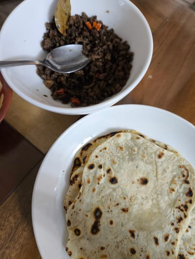 The image shows a close-up view of a meal served on a wooden table. In the upper portion of the frame, a white bowl contains a mound of cooked ground meat, possibly beef or pork, mixed with small pieces of red chili and what appears to be a bay leaf. A silver spoon rests in the bowl, scooping some of the meat. Below the bowl, a white plate holds several folded flatbreads, likely tortillas or similar. The flatbreads are lightly browned with some charred spots, indicating they have been cooked on a grill or pan.

The setting appears to be a casual dining environment, possibly a home or a restaurant. The wooden table surface has a visible grain. The lighting suggests it could be daytime, possibly late morning or afternoon, given the natural light. No people are visible in the frame. The overall impression is a simple, home-style meal. The location context provided is Imus, Philippines, which might suggest the type of cuisine or ingredients used, though specific Filipino dishes are not immediately identifiable without further context. No text is visible in the image.