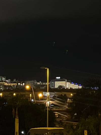 The image depicts a nighttime view of a city, likely Miraverde, Spain, with buildings and streetlights illuminated against a dark sky. In the distance, a large, multi-story building with numerous yellow lights, possibly a hotel or resort, is prominently visible. Several streetlights are present in the foreground and mid-ground, with their yellow glow illuminating the scene. The sky is dark, with three faint, oblong lights that appear to be high in the atmosphere, possibly aircraft or drones. The overall scene suggests a typical urban environment at night, with the added mystery of the unexplained aerial lights.