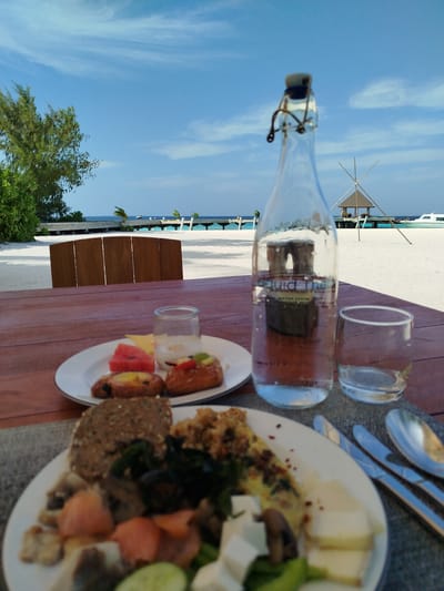 A close-up, eye-level outdoor shot features a wooden table on a sandy beach in Kendhoo, Maldives. The table holds a breakfast spread, including a plate with pastries, fruit, and yogurt, and another plate with a hearty meal of bread, scrambled eggs or frittata, salmon, cheese, and vegetables. A glass bottle of water and a glass sit to the right of the food.
In the background, the turquoise ocean stretches to the horizon under a bright blue sky with scattered clouds. A wooden pier extends into the water, leading to a thatched-roof pavilion. A white boat is visible in the distance on the right. Lush green trees and foliage frame the left side of the scene. The overall atmosphere is serene and tropical.