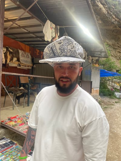 The image shows a man with a beard wearing a white t-shirt and a patterned cap. He is standing in front of a stall that appears to be selling various items, possibly souvenirs, including textiles and small trinkets. The stall is set up outdoors, with a corrugated metal roof supported by metal poles. In the background, there are rocky cliffs, vegetation, and a blue tent. The overall setting suggests a roadside market or tourist area, potentially in a mountainous or natural environment. The man has tattoos on his left arm and is wearing a green wristband.
