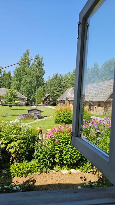 A photograph taken from inside a building looking out through an open window frames a pastoral scene in Sodeliškiai, Lithuania. The window, with its white-painted wooden frame and metal latch, is partially open and angled to the right. The foreground of the image is dominated by a lush garden bed filled with vibrant pink and blue flowers, including phlox, and various green leafy plants. Beyond the garden, a manicured green lawn stretches towards several rustic wooden buildings with thatched roofs. In the middle ground, a traditional well with a stone base and a wooden structure sits on the lawn, accompanied by a tall wooden pole. The background features a dense forest of green trees under a clear blue sky. The overall impression is one of a tranquil rural setting.