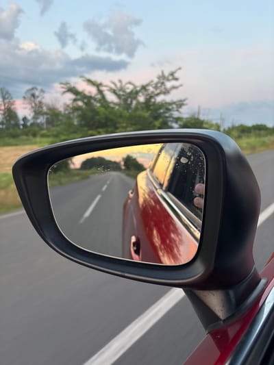 This media file captures a reflection in a car's side mirror. The mirror, mounted on a red car, shows a reflection of the road ahead, the surrounding landscape, and a portion of the car's interior. The scene appears to be set in Bochmanivka, Ukraine, with trees and vegetation visible along the roadside. The sky suggests it is either dawn or dusk, with soft, warm colors of sunset or sunrise reflected in the mirror, blending with the blue of the sky. The road is paved with a white line marking its center, indicating it's a two-way street. In the reflection of the car's interior, part of a hand with fingers visible is seen, possibly the driver's or a passenger's. The overall mood is serene, conveying a sense of movement and contemplation, typical of a journey. The image quality is slightly grainy, adding a touch of artistic texture. There is no visible text in the media file.
