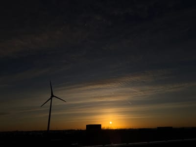 The image displays a panoramic landscape at either sunrise or sunset, identified as Naaldwijk, Netherlands. The sky, occupying the majority of the frame, transitions from a dark blue-grey at the top to vibrant orange and yellow near the horizon where the sun is positioned. Horizontal cloud streaks are visible across the sky, particularly in the middle section. Two distinct white contrails traverse the upper right quadrant of the sky.
The sun appears as a bright, circular orange-yellow orb situated low on the horizon, slightly right of the frame's vertical center. A secondary bright point, possibly a lens flare, is visible above and to the left of the main sun.
The foreground and horizon are rendered in deep silhouette. A tall wind turbine with three blades is prominently positioned in the left half of the image, extending upwards from the horizon line. Multiple indistinct rectangular structures and buildings are silhouetted along the horizon, including a large, dark, box-like building directly below and slightly to the left of the sun. Bare tree branches are discernible in silhouette between this building and the sun. The immediate foreground appears as a dark, flat expanse, with subtle lighter patches potentially indicating snow. No individuals are present in the scene.