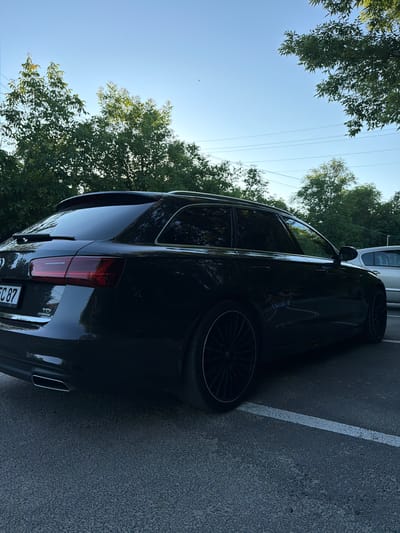 A black Audi A6 Avant is parked on an asphalt surface marked with a white line. The car is viewed from the rear-right quarter, showcasing its taillight, exhaust pipe, and wheel. The vehicle's license plate is partially visible, displaying "EC 87". Above the car, green trees with dense foliage are visible, framing a clear blue sky. Power lines are also present in the sky, indicating an outdoor setting. The lighting suggests it is daytime, likely late afternoon due to the quality of light. There are no people or specific events depicted, just a parked car in an outdoor environment. The scene appears to be in a residential or parking area, given the presence of trees and the paved surface. The overall impression is that of a calm, everyday scene.