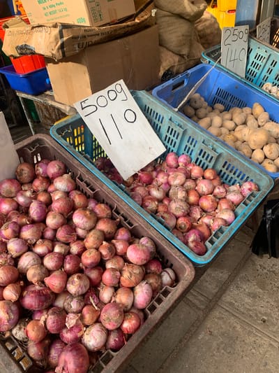 The image shows a close-up, top-down view of a market stall in Kodagoda, Sri Lanka, displaying various produce. In the foreground and middle ground, several blue and brown plastic crates are filled with red onions. The onions vary in size and shade of red and purple, with some showing dry outer skins. Behind the onions, to the left, are crates filled with potatoes. The potatoes are pale yellow to light brown and appear to be of a uniform size.
Several price tags are visible. One white tag in the center, with handwriting in black marker, reads "500g ||". To the left of this, a sign attached to a blue crate has handwritten text in black marker reading "250g 140".
In the background and to the left, sacks made of coarse woven material are piled up, likely containing more produce or other market goods. There are also cardboard boxes, some with labels indicating product information and weight. One visible label on a brown box reads "400g" and "NET WEIGHT 14.4 kg". Another label on a cardboard box indicates "Stacking" and provides a contact number.
The overall scene suggests a vibrant outdoor market, possibly in the morning or afternoon, given the lighting. The focus on fresh produce and the presence of price tags point to a commercial setting where goods are being sold. The ground appears to be concrete or a similar paved surface, common in market areas. The colors are rich and varied, dominated by the earthy tones of the sacks and cardboard, the vibrant reds and purples of the onions, and the lighter tones of the potatoes.