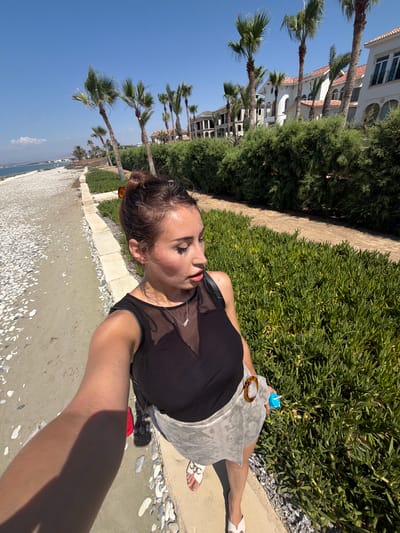 A woman is taking a selfie on a sunny day, standing on a paved walkway next to a beach. She is wearing a black top, a snakeskin-patterned skirt, and white sandals. Her hair is pulled back in a bun, and she is wearing makeup. Behind her, there is a beach with white pebbles, a body of water, a row of palm trees, and a line of white buildings with red-tiled roofs. A dense green hedge runs along the walkway. The scene appears to be a resort area.