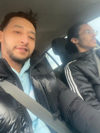 The image shows two individuals, a man in the foreground and a woman in the background, inside what appears to be a vehicle, possibly a car. The image is taken from a low angle, looking upwards towards the occupants.
In the foreground, a man with dark hair and a beard is visible from the chest up. He is wearing a light blue hooded sweatshirt underneath a black puffy jacket. A gray seatbelt crosses diagonally over his chest. He is looking directly at the camera with a slight smile or smirk.
In the background, positioned above and behind the man, a woman is seen lying down or leaning back on a seat. She has dark hair, wears glasses, and her head is resting on a headrest. Her mouth is slightly open, and she appears to be looking upwards and to the right, possibly engaged in conversation or laughing. She is also wearing a black puffy jacket, which features three prominent white stripes on the sleeve, suggestive of an Adidas brand jacket.
The setting is the interior of a vehicle, indicated by the seats, headrests, and a window in the background. Through the window, an overcast or rainy sky is visible, with possible rain streaks on the glass, suggesting an outdoor, daytime scene during cool or wet weather. The interior of the car is primarily dark gray or black.
The activity appears to be a casual selfie taken by the man, capturing himself and the woman in a relaxed moment while traveling or parked in the car. The visible faint white text on the man's light blue hoodie is largely obscured and unreadable.