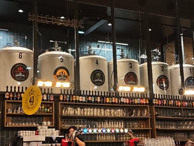 The interior of a bar or brewery features several large, numbered fermentation tanks labeled "La Rouge Brasserie". Shelves behind the bar display a wide variety of bottled beers and glassware. A yellow sign celebrates "40 ANS". A person is partially visible behind the counter near a tap system with multicolored lights. The background wall is tiled.