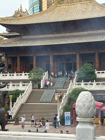 The image depicts a large, ornate Buddhist temple with traditional Chinese architectural elements, including a multi-tiered golden roof with dragon embellishments and intricately carved wooden structures. The temple is accessed via a grand staircase with white marble balustrades. Several people are visible on the stairs and in the plaza area in front of the temple, some appearing to be tourists taking photographs or engaging in conversations. A modern glass skyscraper is visible in the background, suggesting an urban setting. The overall scene conveys a sense of cultural heritage and contemporary life coexisting. The location is identified as the Jing'an Temple in Jing'an District, Shanghai, China.