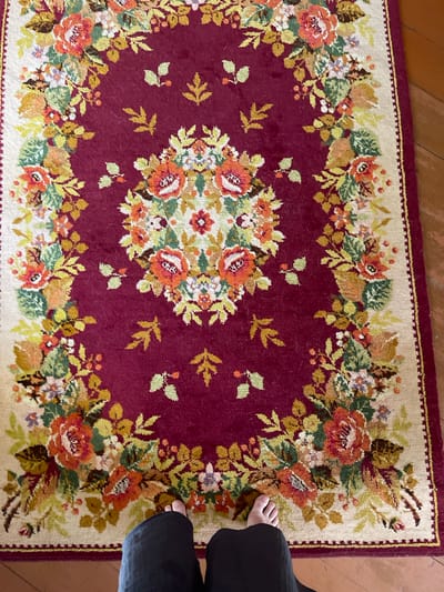 A dark red oriental rug with a floral pattern is laid on a wooden floor. The rug has a cream-colored border and a central floral arrangement composed of various flowers and leaves in shades of orange, yellow, green, and purple. Scattered throughout the red field of the rug are small, stylized leaf motifs. The lower portion of the image shows the bare feet of a person wearing black trousers, standing on the rug. The environment suggests an indoor setting, possibly a home. The location of Калодзежскі сельскі Савет, Belarus, is not visually evident in this image.