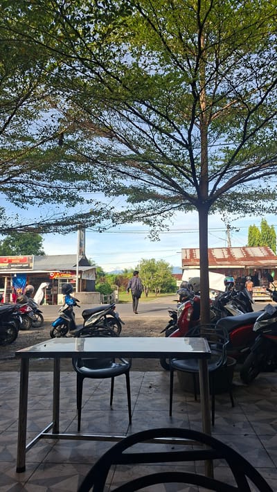 The image shows a street scene with a large tree in the foreground, its branches extending over a sidewalk and street. A metal table with black chairs is set up on a tiled patio. Several motorcycles are parked along the street. A person is walking away from the camera on the sidewalk. In the background, there are buildings, including a shop with a red roof, and lush green trees and mountains in the distance. The sky is clear and blue. The scene appears to be in the city of Keuniree, Indonesia.