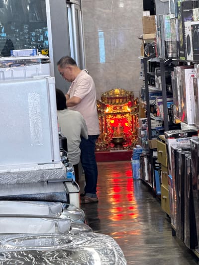 In Ranong, Thailand, a retail store is depicted with a man wearing a pink polo shirt and jeans, and another individual partially obscured from view, both standing near a display of appliances. To the right, shelves are stacked with various electronic items, and further down the aisle, a vibrant, red-lit altar is visible. The floor of the store is a reflective surface, showing a red sheen from the altar's illumination.