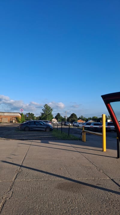 The image depicts an outdoor scene, likely a parking lot, under a clear blue sky with scattered clouds. The perspective is from inside a vehicle with its door open, looking out. In the foreground, the textured surface of a concrete pavement is visible, marked by cracks and shadows cast by an unseen structure. A yellow barrier gate, possibly for a parking entrance or exit, stands prominently to the right, with a black pole extending downwards.
Beyond the barrier, a line of cars is parked along a grassy strip. The cars appear to be a mix of dark colors, with one dark blue or grey sedan clearly visible in the mid-ground. Further in the background, several houses with red-tiled roofs are nestled amongst trees, suggesting a residential or semi-rural setting. Utility poles and wires are also visible, indicating infrastructure.
A red and white circular sign with a red line through it, likely a traffic or parking restriction sign, is visible near the parked cars. A blue parking sign with a white 'P' is also present.
The bright blue sky and the long shadows indicate that it is likely daytime, possibly late afternoon or early morning, given the angle of the shadows. The weather appears to be fair and sunny. The scene evokes a sense of a typical day in a Russian village or small town, Kamyennoye. There are no people visible in the image.