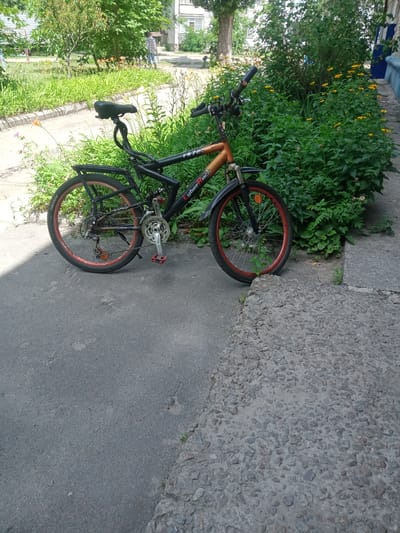 The image shows a black and orange mountain bike parked on a paved area next to a dense green bush with yellow flowers. The bike has a rear rack and red-rimmed wheels. In the background, a person wearing a plaid shirt and blue jeans is walking away from the camera on a path lined with green grass and some orange flowers. Trees are visible behind the person, and a building can be seen in the distance. The ground in the foreground is asphalt, with a section of rough, rocky pavement to the right. The scene appears to be an outdoor area in Vlasivka, Ukraine.