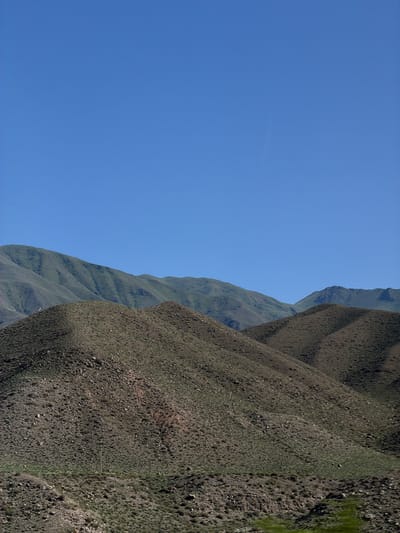 The image displays a vast, clear blue sky on the left, filling roughly two-thirds of the frame. On the right, a steep, rugged mountain slope dominates the scene. The mountain is covered in sparse vegetation and rocks, with visible texture and varied shades of brown and green. The terrain appears arid and uneven, with shadows indicating the contours of the landscape. The sunlight suggests it is daytime. The overall impression is one of natural, remote wilderness, likely in a mountainous region. There are no people, objects, or visible text in the image. The location context provided is Kok Moynok Eki, Kyrgyzstan.
