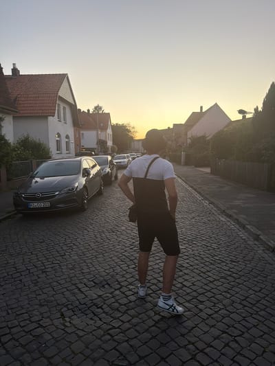 An individual stands on a cobblestone street in Nienburg/Weser, Germany, facing away from the camera. The person wears a dark bucket hat, a white t-shirt with dark horizontal paneling, black shorts, and white athletic shoes. A dark bag is slung across their body. The street is lined with parked cars, including a dark gray Opel Astra station wagon with the license plate "NI CO 201". Residential buildings with red-tiled roofs and white facades are visible on the left side of the street. The scene is illuminated by the warm light of a sunset, casting long shadows and creating a soft glow in the sky. A wooden fence runs along the right side of the street.