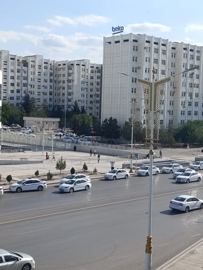 The image shows a street scene in Ashgabat, Turkmenistan on a sunny day. Several white cars, many of which appear to be taxis with green roofs, are parked or driving on the road. In the background, there are large, multi-story white buildings with many windows, typical of Soviet-era architecture. One building prominently features a blue "beko" logo at the top. People are visible walking on the sidewalk and near the buildings. The time of day appears to be afternoon, with the sun casting shadows. The overall scene depicts an urban environment in Turkmenistan, likely during daylight hours.