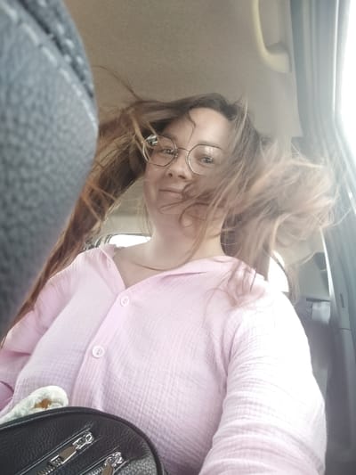 A woman with long brown hair and glasses is in the passenger seat of a vehicle, possibly a car, as indicated by the interior elements visible. Her hair is blowing, suggesting movement or wind. She is wearing a light pink, button-down shirt. In the foreground, a black handbag with silver zippers is visible. The scene appears to be an interior shot taken from a low angle. The location is identified as Блохино, Russia.