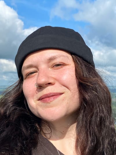 A close-up selfie is shown from the shoulders up, featuring a person wearing a black beanie and a dark-colored shirt. The person has long, dark, wavy hair and is looking directly at the camera. Their face is slightly tilted, and they have a silver nose ring. The background is a bright blue sky with scattered white clouds, suggesting an outdoor setting on a sunny day. The image does not provide any information about the location in Andreevka, Russia.