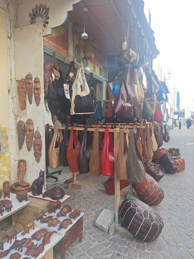 This image captures a street scene in Essaouira, Morocco, likely in the medina given the traditional architecture and the presence of artisan shops. The foreground is dominated by a shop displaying a variety of leather goods, primarily handbags and poufs, suspended from wooden racks or arranged on the cobblestone street. Wooden carved masks and small animal figurines are also prominently displayed on shelves to the left of the shop entrance. The shop itself appears to be a small, open-fronted establishment with a weathered facade and an awning providing shade.
In the background, a narrow street stretches into the distance, lined with buildings that suggest a historic urban environment. The light is bright, indicating it is likely daytime. A few people are visible in the background, walking along the street, suggesting ongoing commercial activity. The overall impression is of a bustling marketplace, typical of Moroccan souks, showcasing local craftsmanship and trade.