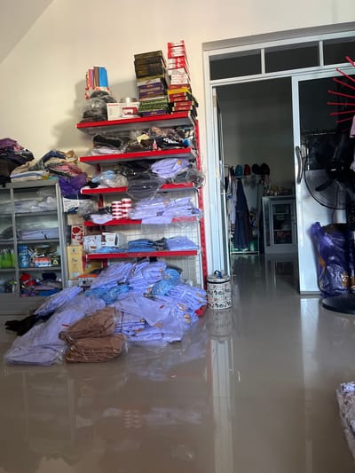 The image depicts the interior of a retail store or storage space in Unnyi, Indonesia, filled with merchandise, primarily clothing. The scene is cluttered and organized in a functional rather than aesthetic manner.

Dominating the foreground and midground are stacks of white and light blue folded garments, many of which appear to be school uniforms or similar attire, encased in plastic packaging. Some bundles are tied with string. To the left, a gray metal shelving unit is filled with various items, including boxes, plastic containers, and rolled fabrics.

To the right, red metal shelves are stacked high with more packaged clothing, boxes of what appear to be gifts or packaged goods, and some household items like a red and white striped canister and a white appliance box. Above these shelves, more boxes are stacked precariously. A doorway leads into a dimly lit interior space where additional shelves and items are visible, including what looks like a small cabinet.

In the far right of the image, a tall black standing fan is positioned, partially obscuring a large purple plastic bag. The floor is tiled and highly reflective, showing the clutter of the space. The lighting appears to be artificial, creating a somewhat dim and utilitarian atmosphere. There are no people visible in the image. The overall impression is of a busy retail environment where stock is being managed.