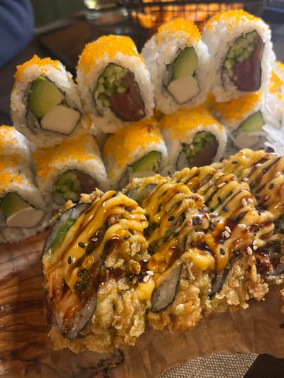A close-up shot displays two types of sushi rolls arranged on a wooden surface. The left side showcases sushi rolls with orange roe coating, filled with ingredients like tuna, cucumber, avocado, and imitation crab. Adjacent to these are fried sushi rolls drizzled with a creamy yellow sauce and a darker, possibly soy-based, glaze, sprinkled with sesame seeds. The sushi appears freshly prepared and visually appealing.