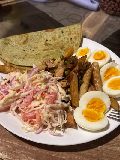 The image shows a white plate of food, containing a green tortilla, penne pasta with peas and chicken, hard-boiled eggs cut in half, and a salad of shredded cabbage, red onion, and tomatoes. A fork rests on the plate. The plate sits on a wooden surface. To the upper left, there's a glimpse of what appears to be a container of peanuts and a white object. The location cannot be determined, and the image does not include any people, actions, or interactions.