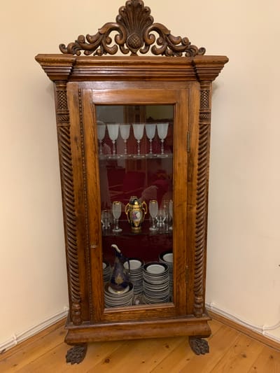 This is a photograph of a vintage wooden display cabinet filled with various items. The cabinet has a dark wooden finish with ornate carvings on the top corners and sides. The front of the cabinet features a glass door, through which the contents are visible.
Inside the cabinet, on the left side, are several rows of clear stemmed glasses, possibly wine glasses, with frosted bowls. On the right side, there are stacks of plates with a dark rim and a white center, and a decorative blue pitcher with gold embellishments is nestled amongst them. A small, ornate golden vase with painted details is also visible. The interior of the cabinet appears to be lined with a dark red fabric.
The cabinet is positioned in what appears to be a domestic setting, against a plain light-colored wall. To the right of the cabinet, the wall transitions to a diagonally paneled wooden surface, suggesting a corner of a room. The lighting is even, indicating it was taken indoors with artificial light, and there are no indications of the time of day or weather. No people are visible in the image, and no text is present. The overall impression is one of an antique piece of furniture displaying a collection of tableware and decorative items.