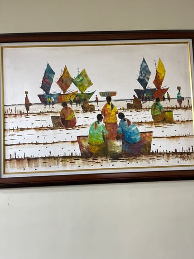 The painting depicts a waterfront scene with several boats and people. The boats are large with colorful sails in shades of blue, yellow, and green. The people are depicted as stylized figures, some carrying baskets on their heads, while others are gathered around containers, possibly engaged in a market activity or preparing goods. The foreground is rendered with textured brushstrokes suggesting water or muddy ground with wooden posts. The overall style is impressionistic, with bold colors and simplified forms. The painting is framed in dark wood with a gold inner trim. The location is identified as Quezon City, Philippines.