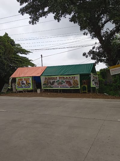 A roadside food stall, named "Arum Ndalu," is set up with orange and green tarps. The stall advertises "Goreng Bakar" (fried and grilled) and "Pecel Lele" (catfish). Banners feature images of chicken and catfish dishes. The stall is situated beside a road, with trees and an overcast sky visible in the background. A small sign with text is partially visible near the right side of the stall.