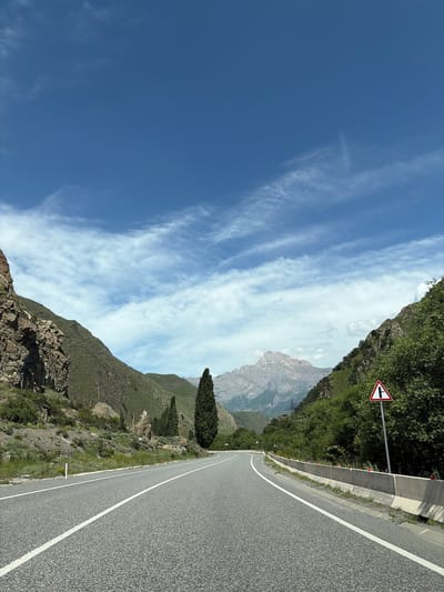 The image depicts a scenic mountain road in Mizur, Russia. The road is paved and has white lane markings. It curves to the right, leading towards a distant mountain range. The mountains on either side of the road are covered in green vegetation and rocks. The sky is blue with scattered white clouds. There is a traffic sign on the right side of the road indicating a fork in the road. The scene appears to be a clear, sunny day. No people or animals are visible in the image.
