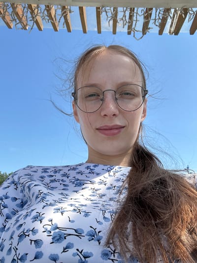 A young woman with long brown hair and glasses is taking a selfie outdoors. She is wearing a white dress with a blue floral pattern. Her hair is windswept and partially covers her face. The background is a clear blue sky, with a wooden structure adorned with string lights visible at the top of the frame. A sliver of greenery is visible on the far left.