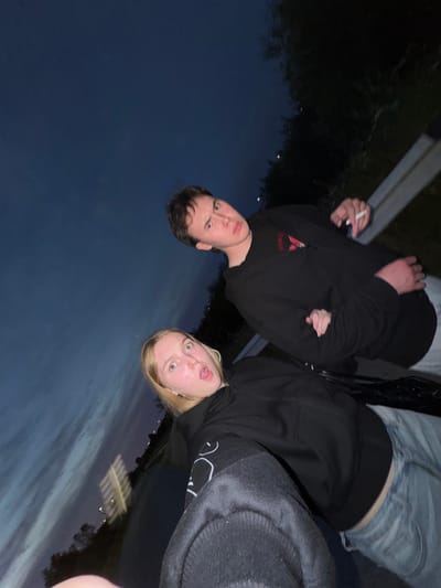 The image depicts two young individuals, a male and a female, at dusk, possibly in the vicinity of the village of Ryabinino, Russia. The male is positioned in the upper right, looking towards the camera with a slightly surprised expression. He is wearing a black hooded sweatshirt and holding a lit cigarette in his right hand. The female is in the lower left, also facing the camera with her mouth open in an exaggerated expression. She is wearing a black hooded sweatshirt and light-colored jeans. They appear to be leaning against a dark, possibly metallic surface, like a car. The background shows a dimly lit sky with faint clouds and silhouetted trees and structures in the distance. The overall impression is a candid, informal snapshot taken outdoors during twilight.