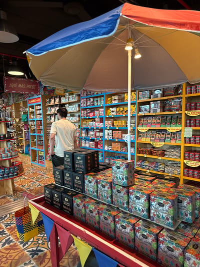 An indoor market scene in the Area Metropolitana Centro Occidente (AMCO), Colombia, is observed from a slightly elevated perspective. A single male individual, seen from behind, with dark hair, wearing a light tan t-shirt and dark pants, stands facing a wall of merchandise shelves on the left side of the frame. In the center foreground, a large, open market umbrella features blue, beige, red, and yellow colored sections, with two small lights attached to its central pole. Beneath the umbrella, a red, boat-shaped display stand, adorned with small red, yellow, and blue pennants, exhibits multiple stacks of rectangular "CAFE COFFEE" tins. Some tins are solid black with gold lettering, while others display a vibrant, multicolored, leaf-like pattern.
Behind the umbrella and the individual, extensive shelving units, colored in bright blue and yellow, line the walls, filled with a diverse array of packaged food products, including coffee, snacks, and numerous chocolate bars with visible pricing, such as "3X $59.900" and "3X $99.900 / 1X $44.900." A hanging wooden sign on the left reads "¿QUÉ MÁS PUES?" and another, "EL MARKET COLOMBIA." The floor features an intricate, multicolored geometric tile pattern. A woven, multicolored basket rests on the floor to the left of the boat display. Overhead, exposed ceiling structures and recessed lighting fixtures are visible.