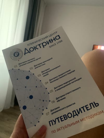 The image shows a close-up of a person's hand holding a brochure or booklet. The brochure has a white background with blue text and graphics. The text is in Russian and appears to be a guide or a set of principles related to a clinical center. Some of the visible text includes "КЛИНИЧЕСКИЙ ЦЕНТР" (Clinical Center), "Доктрина" (Doctrine), and "ПУТЕВОДИТЕЛЬ" (Guidebook). The graphics include a stylized brain and a network of interconnected dots and lines. The lighting suggests it might be daytime, and the setting appears to be indoors. The hand holding the booklet has manicured nails with pink polish. The orientation of the booklet suggests it is being held up and viewed from above, possibly for a photograph or to share information. There are no obvious clues about the specific location within Poritsy, Russia, beyond the text on the booklet itself.