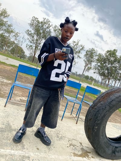 A full shot shows a Black person standing outdoors and looking at a phone. They are wearing a black long-sleeved jersey with white stripes on the sleeves and white lettering and the number 29 on the front. They are also wearing dark, wide-leg jeans, grey socks, and shiny black platform loafers with silver buckles. The person's hair is styled in several small, round buns. They are standing on a concrete surface, with two blue metal chairs behind them. A large, worn tire is visible to the right of the frame. The background shows a chain-link fence, trees, and a cloudy sky. The scene appears to be an outdoor recreational or school area in Zawan, Nigeria.