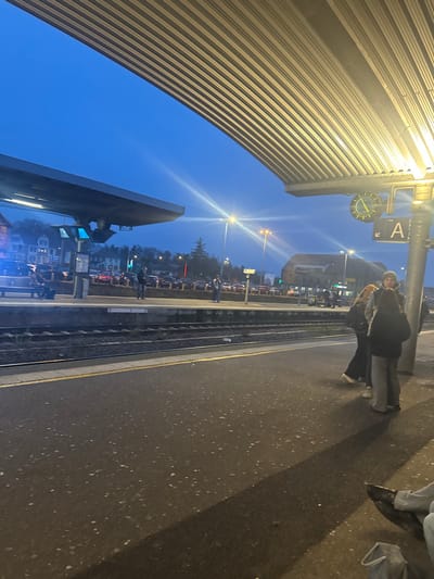 The image depicts a train station platform at dusk or night, brightly illuminated by artificial lights. A large, curved canopy with a corrugated texture extends over the left side of the platform. On the platform, two individuals are standing near a metal support pillar, behind which a white sign with a large black letter "A" and a round analog clock are attached. The clock face is light with dark hands and markers. The lower legs and feet of another person are visible in the foreground on the right.

Train tracks run parallel to the platform, with a smaller platform structure visible on the opposite side. Beyond the tracks, a townscape is visible against the dark blue twilight sky. This background includes buildings, numerous parked vehicles, and trees, all lit by streetlights that create prominent lens flares in the scene. The overall impression is of people waiting at a train station on a clear evening.